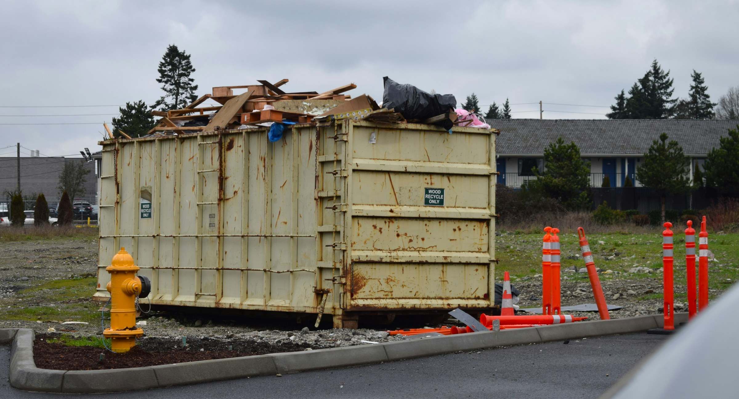 A roll-off can loaded with scrap wood next to a fire hydrant and orange posts